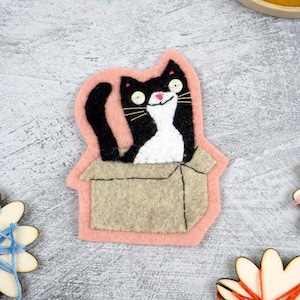 May include: A felt applique featuring a black and white cat sitting inside a tan cardboard box. The cat has white button eyes, a pink stitched nose, and long whiskers. The entire design is outlined in a soft pink felt border on a gray surface.