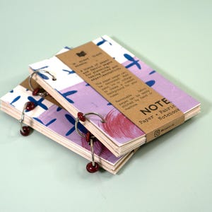 Puede incluir: Dos cuadernos apilables con un diseño abstracto colorido en la portada. Los cuadernos están encuadernados con anillas metálicas y tienen una etiqueta de papel marrón que dice "NOTE Paper + Fabric Notebook".