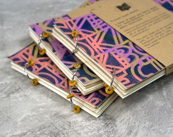 May include: A stack of three small, colorful sketchbooks with a geometric pattern on the cover. The sketchbooks are bound with metal rings and have yellow plastic beads on the rings. The top sketchbook has a brown paper label with the text "Sketch Book" on it.