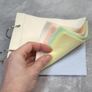 May include: A felt book with a cream cover and silver binder rings. The pages are pastel shades of pink, green, and yellow. The book is open, displaying the colourful pages. The felt squares are soft and inviting.