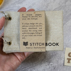 May include: A fabric sketchbook with a linen cover, held open by metal rings. The cover has a brown paper label with text that reads "STITCH BOOK Fabric Sketchbook". The sketchbook has 8 blank pages ready to stitch.