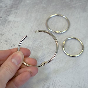 May include: Three silver metal binder rings. The largest ring is open and being held by a hand. The rings are used to hold papers together.
