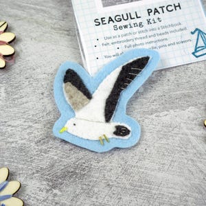 May include: A felt seagull patch featuring a white body, black wing tips, and a yellow beak, set against a light blue background. The patch is positioned next to a sewing kit package labelled SEAGULL PATCH Sewing Kit with instructions.