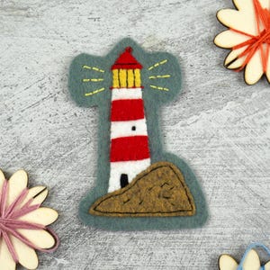 May include: A felt applique patch featuring a lighthouse with horizontal red and white stripes, a yellow lantern room, and a brown rocky base, all set against a dark grey felt background with yellow embroidered light beams radiating outward.