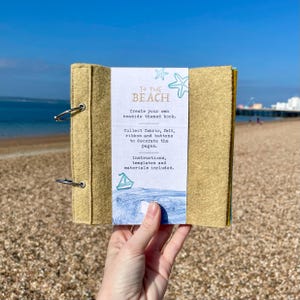 Peut inclure: Un livre fait à la main, recouvert de feutre, avec des anneaux de reliure en métal. La couverture est de couleur beige clair, et une étiquette blanche indique "TO THE BEACH" avec des instructions pour créer un livre sur le thème de la mer.