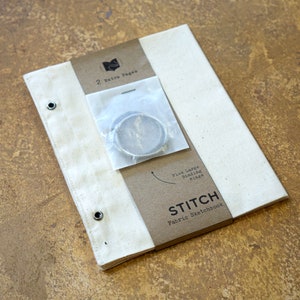 May include: A white canvas sketchbook with the text "STITCH Fabric Sketchbook" printed on the front cover. The sketchbook has two extra pages and plus large binding rings.