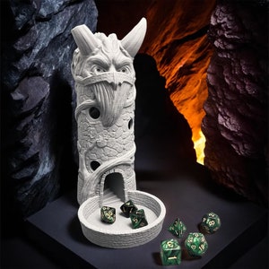 Pode incluir: Uma torre de dados cinza claro com um design detalhado de rosto demoníaco, chifres e uma textura semelhante à pedra. A torre tem uma pequena porta arqueada e uma bandeja circular. Vários dados verdes e pretos estão espalhados ao redor da bandeja.
