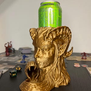 May include: A gold-colored, 3D-printed can holder featuring a detailed female face with horns and a snake. A green and silver can of "KARNITYNA" energy drink sits in the holder. The holder is on a table with game pieces and dice.