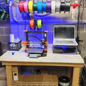 May include: A 3D printer with a spool of filament on top, a laptop computer, and a workbench with boxes of filament underneath. The printer is orange and black, and the laptop is silver. The workbench is made of wood and has a light brown finish.