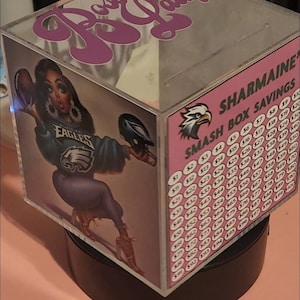 Może przedstawiać: Przezroczysta akrylowa skrzynka z różową i białą etykietą z napisem "SHARMAINE'S SMASH BOX SAVINGS". Skrzynka ma obrotową podstawę i zawiera rysunkową ilustrację kobiety w niebiesko-zielonej koszulce Philadelphia Eagles. Skrzynka ma szczelinę na górze do wrzucania pieniędzy.