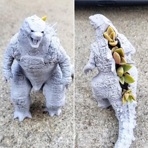 Może przedstawiać: Szary, wydrukowany w 3D doniczka Godzilla z sukulentami rosnącymi z jego pleców. Doniczka stoi na szarej betonowej powierzchni.