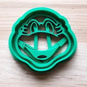 May include: A bright green plastic cookie cutter in the shape of a cartoon frog's face. The cutter has detailed features including eyes, a nose, and a smiling mouth. The cutter is designed for making frog-shaped cookies.