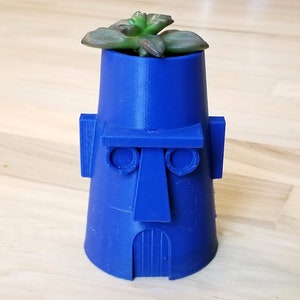 Puede incluir: Una maceta azul, impresa en 3D, con forma de estatua Moai, con una pequeña planta suculenta en la parte superior. La maceta tiene una cara detallada con ojos, nariz y boca, y una puerta en la base.