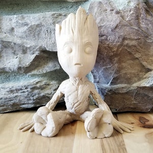 Può includere: Una statuetta di legno marrone chiaro raffigurante un personaggio dall'aspetto arboreo, seduto con le gambe distese. La statuetta ha una consistenza dettagliata ed è ambientata su uno sfondo di muro di pietra.