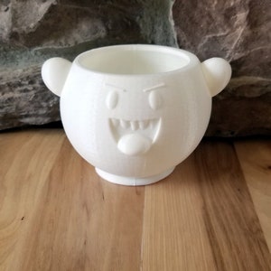 May include: A white, 3D-printed planter in the shape of a cartoon ghost, featuring a wide, toothy grin and a protruding tongue. The planter has two small, rounded ears and a circular opening. It is placed on a wooden surface.