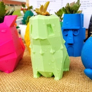 Puede incluir: Una colección de macetas coloridas de forma geométrica, cada una con una pequeña planta suculenta. Las macetas tienen varias formas, incluyendo un perro, un personaje con una cara y una forma redonda con una cara sonriente. Los colores incluyen rosa, verde, azul y amarillo.