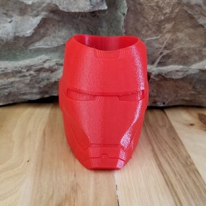 Puede incluir: Un macetero rojo brillante impreso en 3D con forma de casco de Iron Man. El macetero tiene una superficie texturizada y presenta el icónico diseño de la cara con aberturas para los ojos y la boca. Está sobre una superficie de madera.