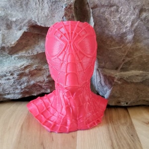 Puede incluir: Busto rojo vibrante de Spider-Man, que muestra la máscara y el cuello del personaje. La escultura impresa en 3D se encuentra frente a un fondo de piedra texturizada y una superficie de madera, destacando el diseño detallado y el color.