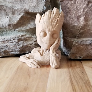 Pode incluir: Uma figura de madeira do personagem Groot, dos Guardiões da Galáxia, com um rosto sorridente e uma mão apoiada no queixo. A figura está sentada sobre uma superfície de madeira.