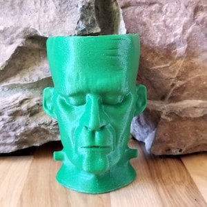 Könnte beinhalten: Ein grüner 3D-gedruckter Pflanztopf in Form von Frankensteins Kopf. Der Topf hat ein detailliertes Gesicht mit geschlossenen Augen und einem neutralen Ausdruck.