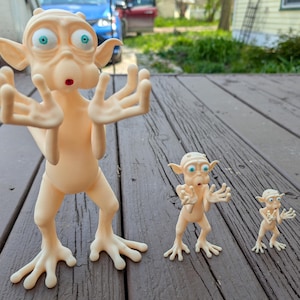 May include: Three light beige alien figurines of varying sizes, each featuring large pointed ears, wide blue eyes, a small red mouth, and four-fingered hands raised in a gesture of surprise, standing upright on a dark wooden surface.