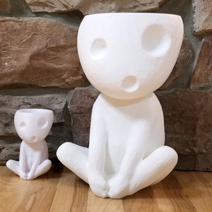Puede incluir: Dos macetas de cerámica blanca con forma de personaje de un videojuego. La maceta más grande está sentada en un suelo de madera con una pared de piedra en el fondo. La maceta más pequeña está delante de la más grande.