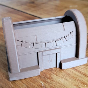Puede incluir: Un modelo impreso en 3D de un pequeño edificio con un techo curvo y una puerta. El edificio tiene una hilera de banderas colgando sobre la puerta.