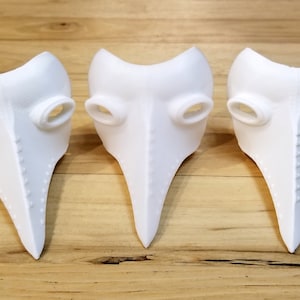 Könnte beinhalten: Drei weiße Pestdoktormasken mit langen Schnabelnasen und ovalen Augenlöchern werden auf einer Holzoberfläche gezeigt. Die Masken haben eine strukturierte Oberfläche und bestehen aus einem festen, undurchsichtigen Material. Die Masken sind ein historisches Kostüm.