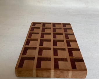 May include: A rectangular wooden cutting board with a geometric pattern of light and dark wood. The board has a light brown background with a grid of darker brown squares and purple squares.