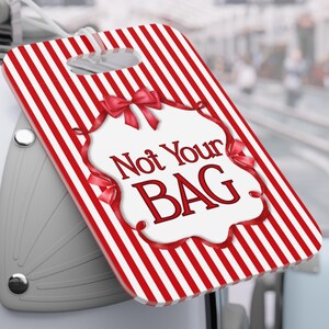 May include: A red and white striped luggage tag with the text "Not Your BAG" in a decorative frame. The tag is attached to a white suitcase, ideal for travel and identifying luggage.