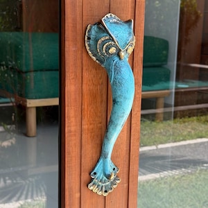 May include: A vertical door handle shaped like an owl with a turquoise patina finish and gold-toned accents. The handle features a detailed owl head at the top and a flared, feather-like base, mounted onto a vertical wooden door frame.