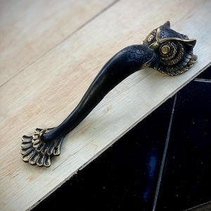 May include: A black and gold owl-shaped door handle with intricate details. The handle is made of metal and has a long, curved design. The owl's eyes are large and round, and its feathers are detailed.