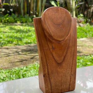 May include: A wooden jewelry display stand with a rounded top and a rectangular base. The stand is made of a light brown wood and is designed to hold necklaces and pendants. The stand is placed on a white surface with a green background.