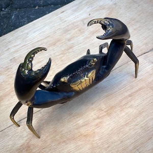 May include: A detailed bronze crab sculpture with a dark finish and gold accents on the claws and face. The crab is posed with its claws raised, showcasing intricate detailing. The sculpture is placed on a light wooden surface.