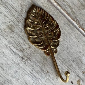 May include: A gold-colored, leaf-shaped wall hook. The hook is made of metal and has a detailed, textured design. The hook is mounted on a weathered, white-painted wooden surface.