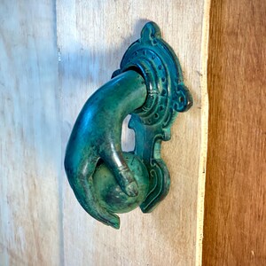 Puede incluir: Un aldaba de puerta de color verdín en forma de mano que sostiene una esfera. La aldaba está montada en una puerta de madera de color claro. La mano y la placa trasera decorativa presentan detalles intrincados, lo que sugiere un diseño antiguo o vintage.
