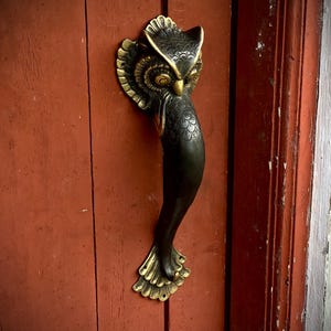 May include: A black and gold owl door handle with intricate details. The handle is attached to a red wooden door.