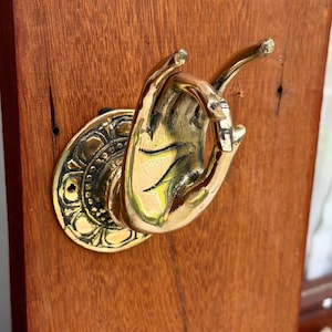 May include: A polished brass wall hook shaped like a hand with fingers positioned in a mudra gesture. The hand is attached to a circular, ornate brass base featuring a decorative floral pattern, mounted onto a vertical piece of reddish-brown wood.