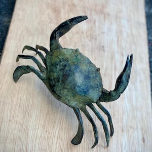 May include: A detailed bronze crab sculpture with a weathered, verdigris finish. The crab has multiple legs, claws, and a textured shell. The sculpture is placed on a light-colored wooden surface.