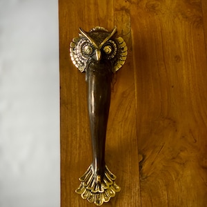 May include: A gold owl door handle with a detailed design. The handle is attached to a wooden door.