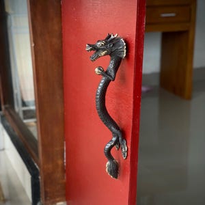 May include: A black dragon-shaped door handle with gold accents is mounted on a red door. The dragon's head is at the top, and its body curves down the door. The handle is made of metal and has intricate details.