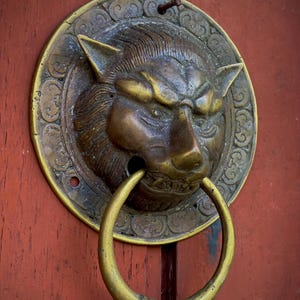 May include: A detailed, antique brass door knocker featuring a lion's head with a ring handle. The knocker is mounted on a weathered, reddish-brown wooden door. The lion's head has intricate details and a fierce expression.
