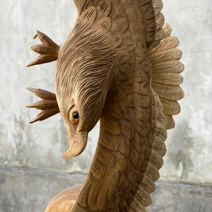 Op de afbeelding: Een houten beeldhouwwerk van een adelaar met zijn vleugels wijd uitgespreid. De adelaar is realistisch gebeeldhouwd, met gedetailleerde veren en een scherpe snavel. Het hout is lichtbruin van kleur en de ogen van de adelaar zijn donkerbruin geverfd.