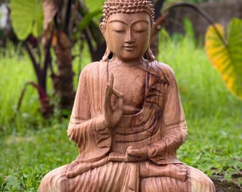 May include: A hand-carved wooden Buddha statue in a seated lotus position. The figure features a textured, pointed ushnisha, closed eyes, and a serene expression. The right hand is raised in a mudra gesture, while the left rests on the lap.