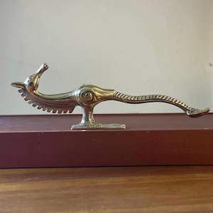 May include: A polished brass figurine of a stylized mythical creature with a long, slender neck, a serrated crest along the underside, a curved body, and a long, textured tail, mounted on a rectangular base with a flat, stable bottom.