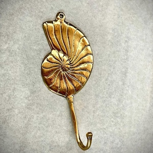 May include: A polished gold-toned metal wall hook shaped like a nautilus shell. The decorative piece features a spiraling, ribbed shell design with a small circular mounting hole at the top and a curved hook extending from the base for hanging items.
