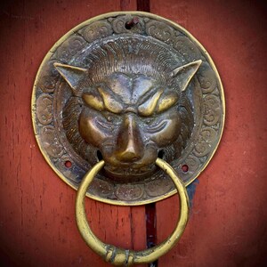 May include: A circular brass door knocker featuring a sculpted lion head with a prominent mane and a smooth, round ring held in its mouth. The metal has a weathered, aged patina with dark highlights, mounted on a textured, deep red wooden surface.