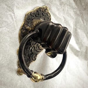Puede incluir: Aldaba de puerta ornamentada con diseño de puño. La aldaba presenta un puño de bronce oscuro y un anillo negro, montado sobre una placa trasera de latón decorativa con patrones detallados. El diseño es vintage y decorativo.
