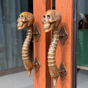 May include: A pair of brass door handles shaped like human skulls attached to spinal columns. Each handle features a detailed cranium at the top, a segmented vertebral column forming the grip, and a pointed base, mounted on ornate plates.