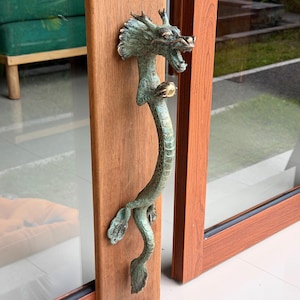 May include: A vertical door handle shaped like a dragon with a weathered green patina finish. The dragon features a detailed head with open jaws, a long serpentine body with textured scales, and a small gold-toned sphere held in its claw.